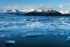 La lagune glaciaire de Jokulsarlon est gelée en surface, immobilisant les plaques de glace, le vent qui souffle fort irise la surface du lac au bord des rives.